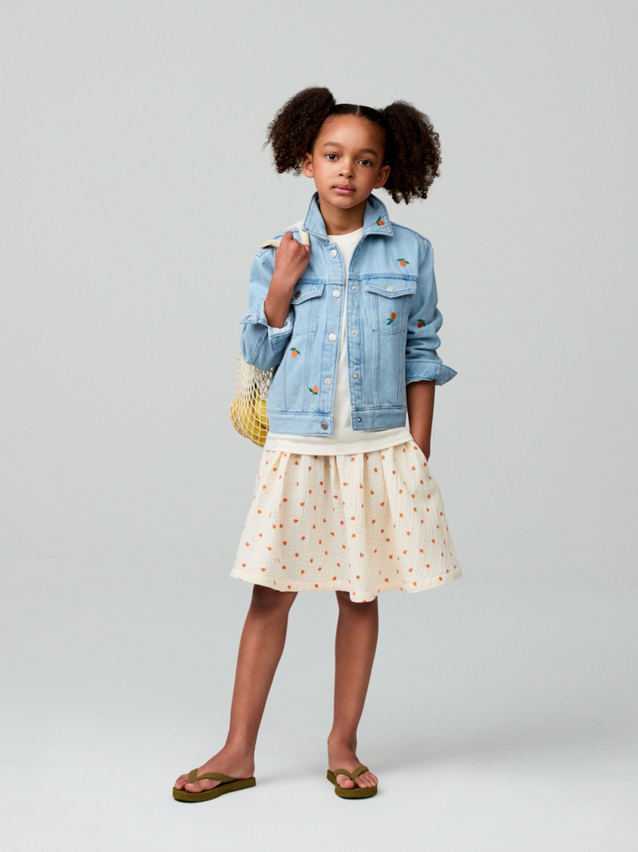 A young girl in a denim jacket and polka dot skirt stands with a yellow net bag, wearing flip-flops.