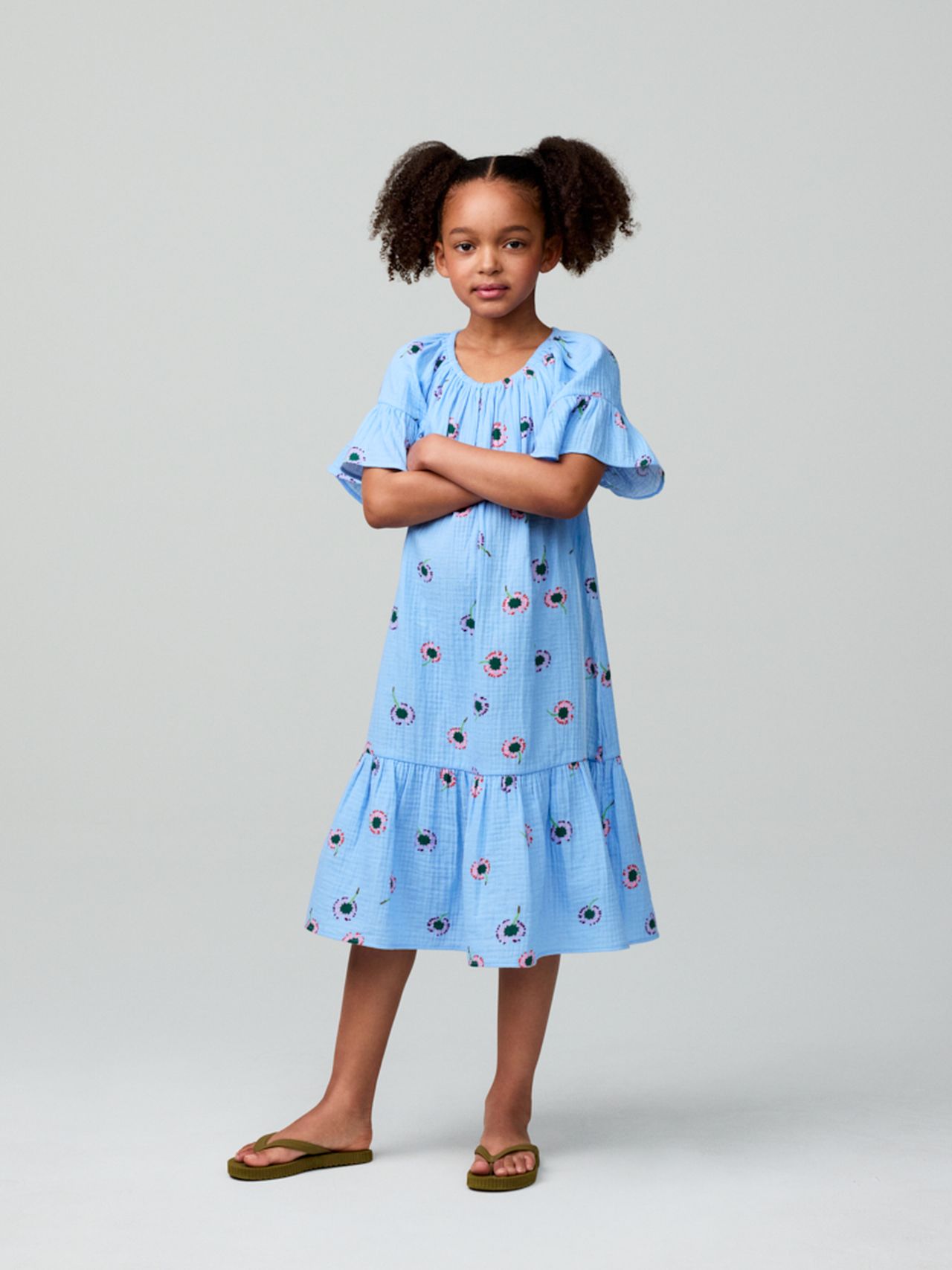 Young girl in a blue dress with floral patterns stands confidently with arms crossed, wearing sandals, against a plain background.