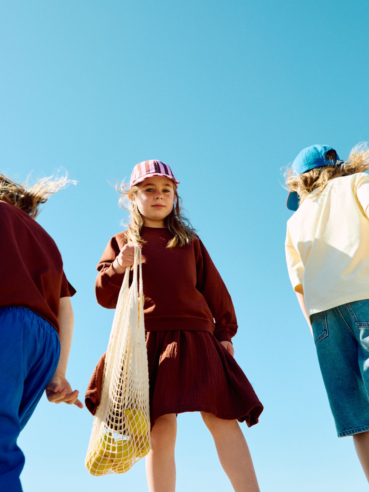 Three children in colorful outfits and caps stand under a clear blue sky. One holds a net bag with yellow items.