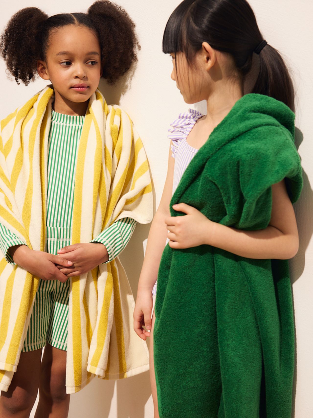 Two girls standing against a wall, wrapped in towels—one in yellow stripes, the other in solid green—engaging in conversation.