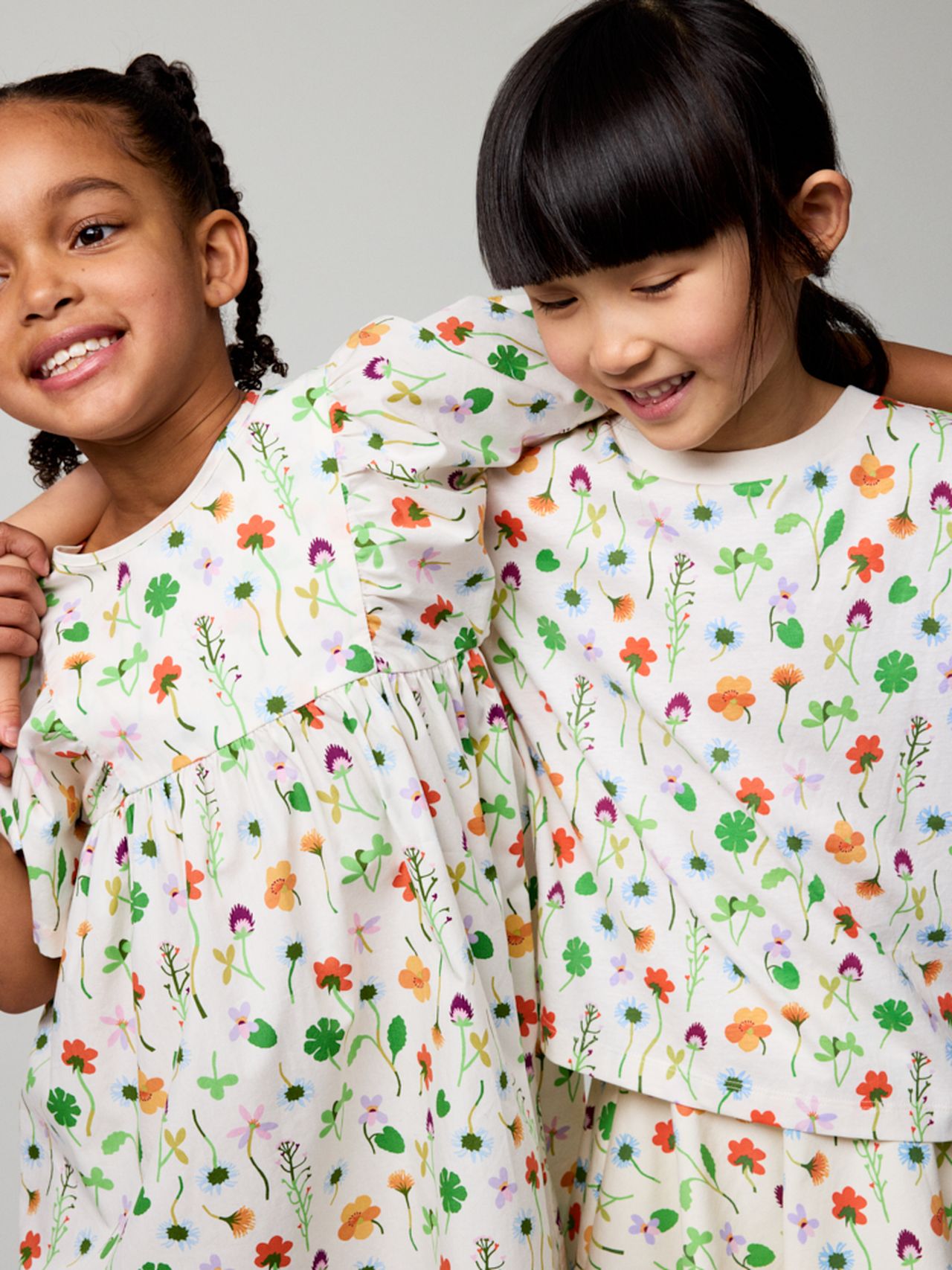 Two young girls smiling, wearing matching floral-patterned dresses, with one girl's arm around the other's shoulder against a plain background.
