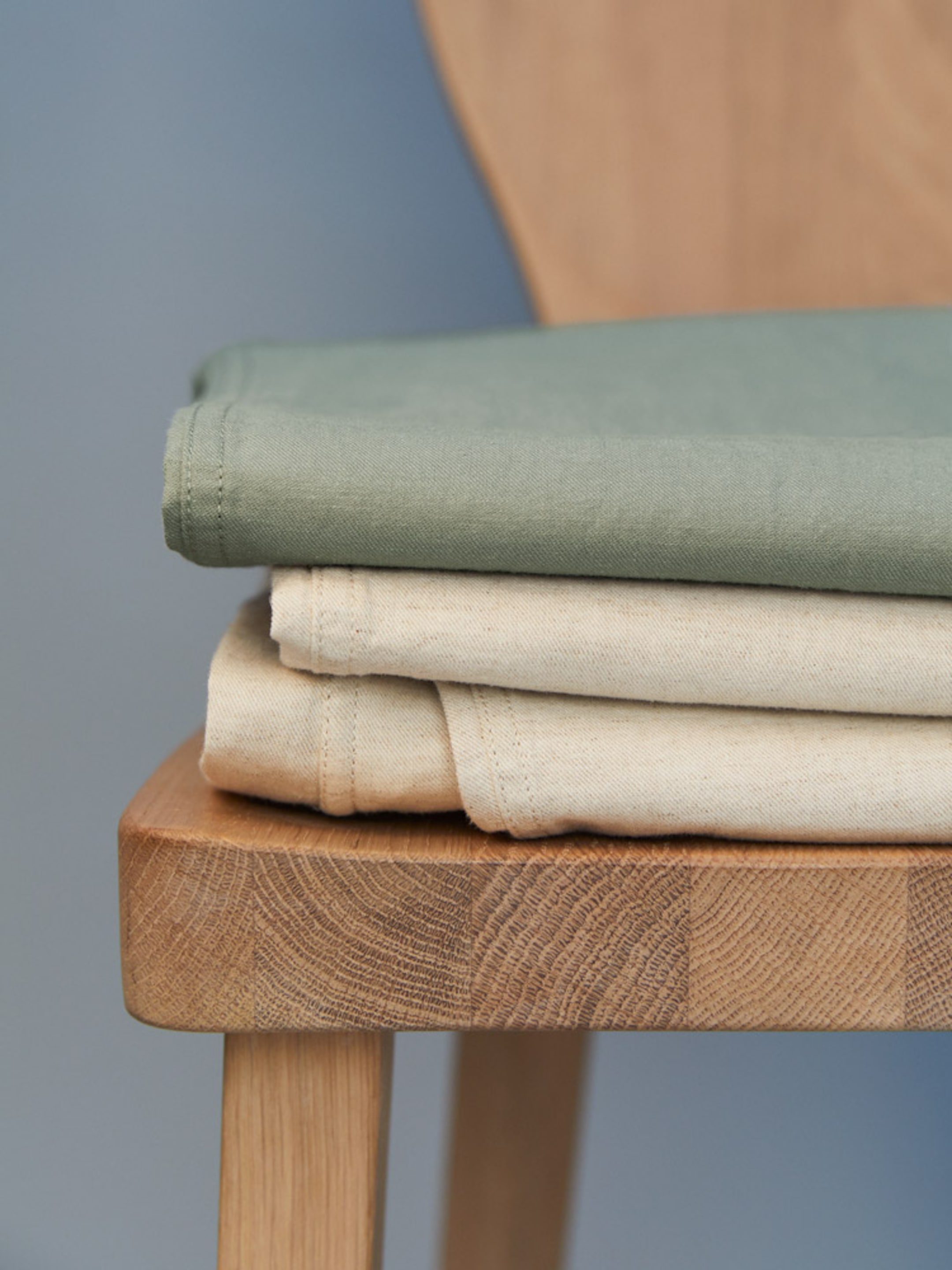 Folded green and beige fabric neatly stacked on a wooden chair, set against a soft blue background.