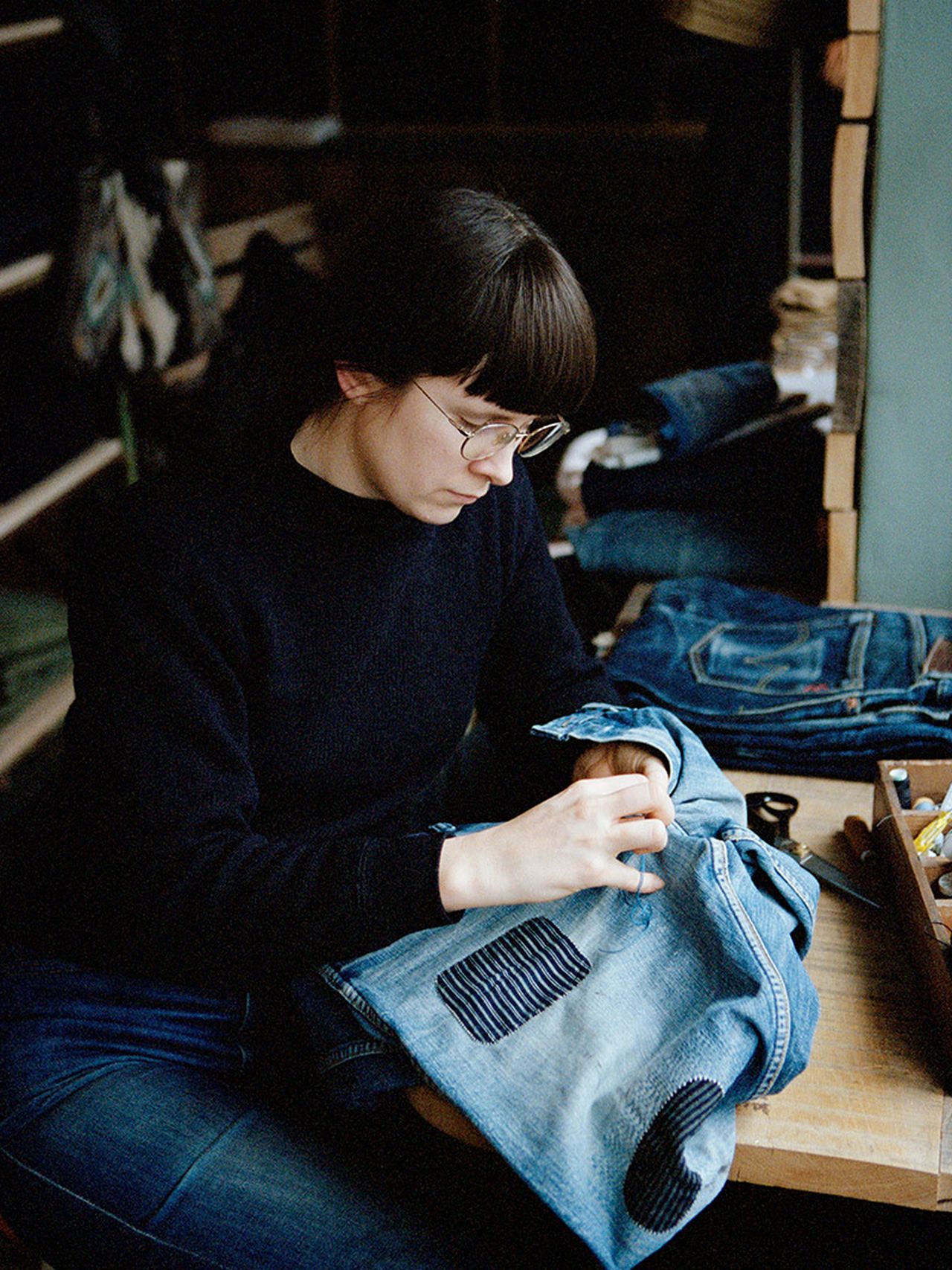 Person sewing patches onto denim jeans at a wooden table, surrounded by sewing tools and materials.