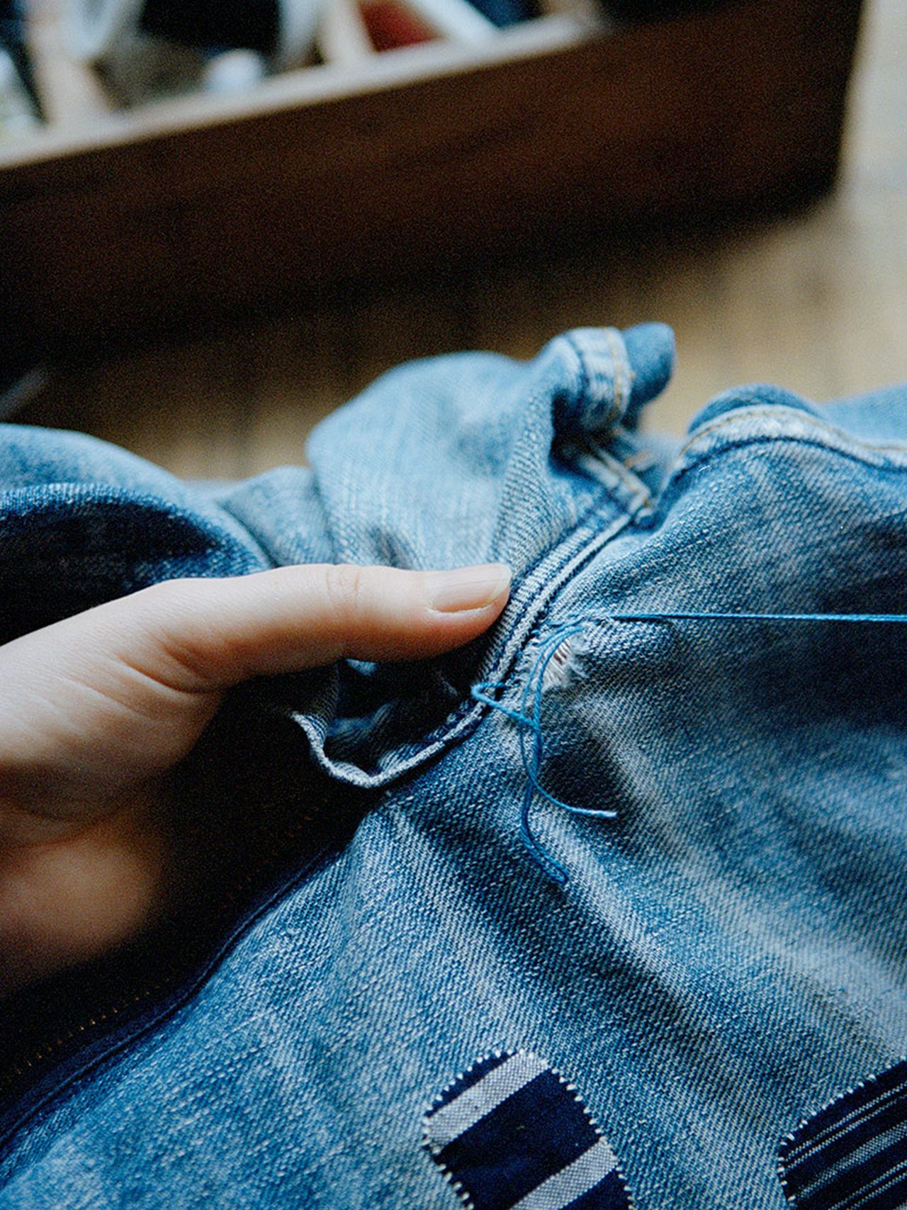 A hand sewing a patch onto blue denim jeans with blue thread, showcasing a close-up of the stitching process.