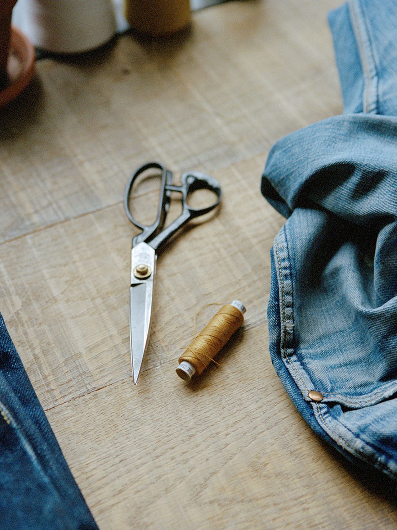 Scissors and a spool of brown thread on a wooden table next to blue denim fabric.