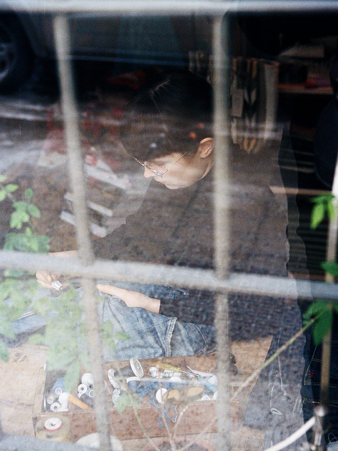 Person sitting indoors, viewed through a window, focused on crafting with tools and materials on the floor.