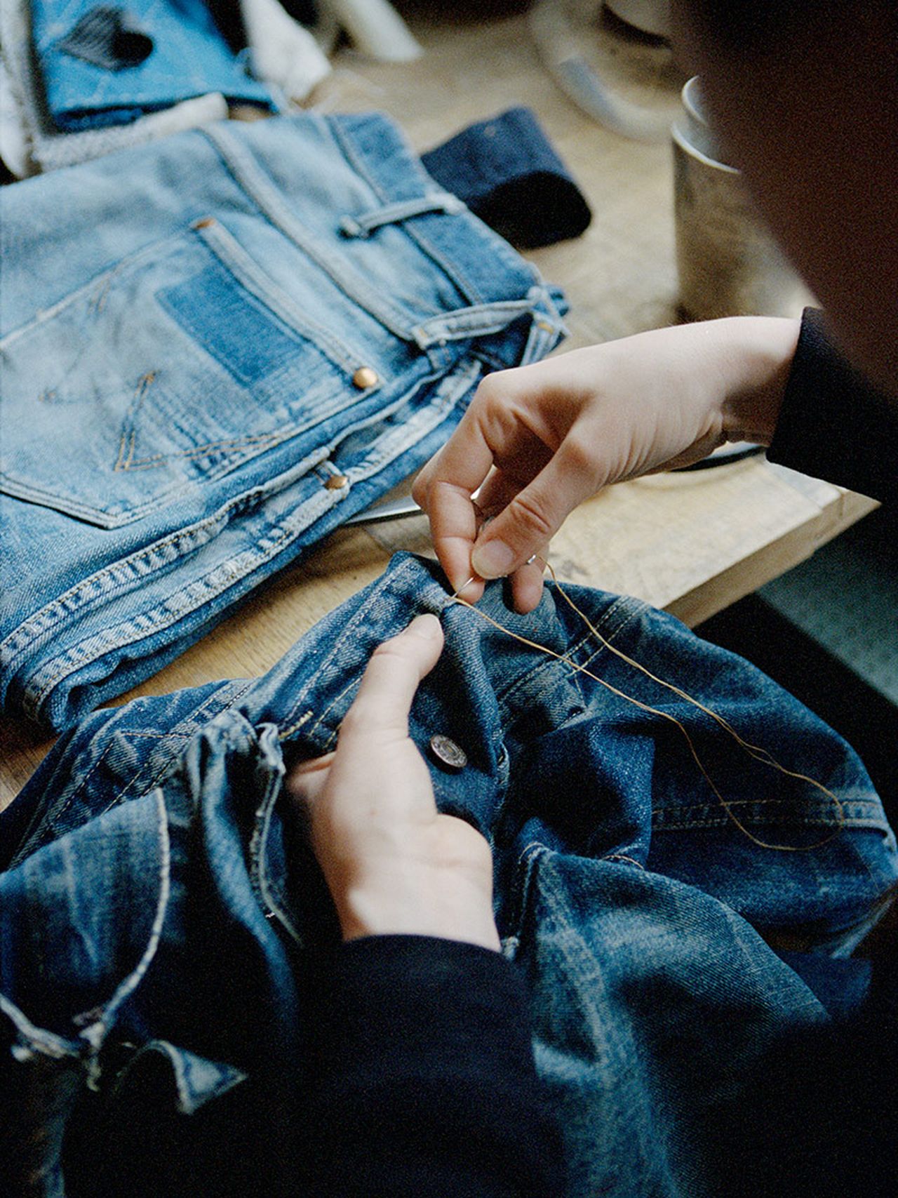 Person sewing a button on blue jeans with a needle and thread, surrounded by denim fabric on a wooden surface.
