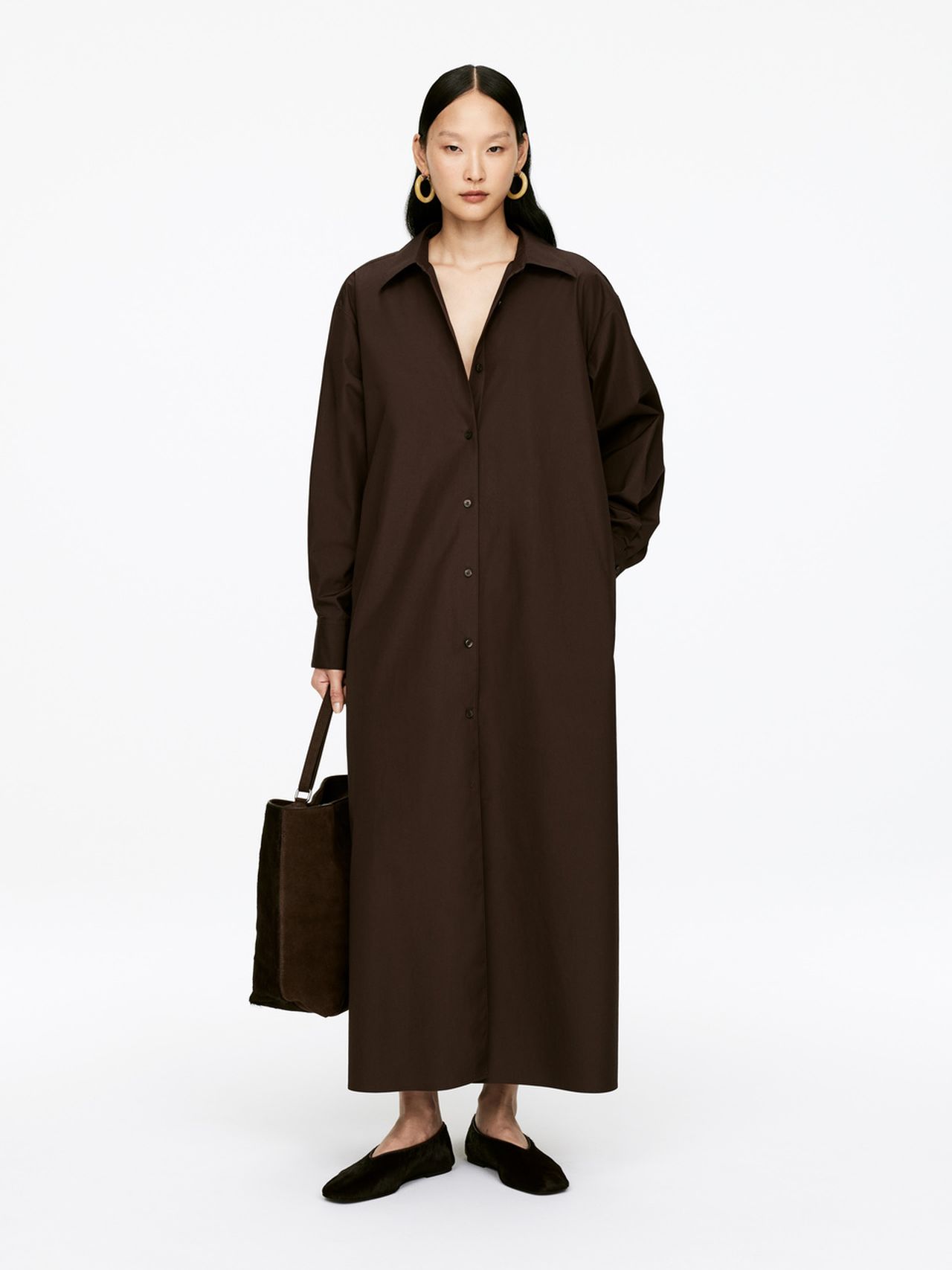 Woman in a long brown button-up dress, holding a brown bag, wearing black shoes, standing against a white background.