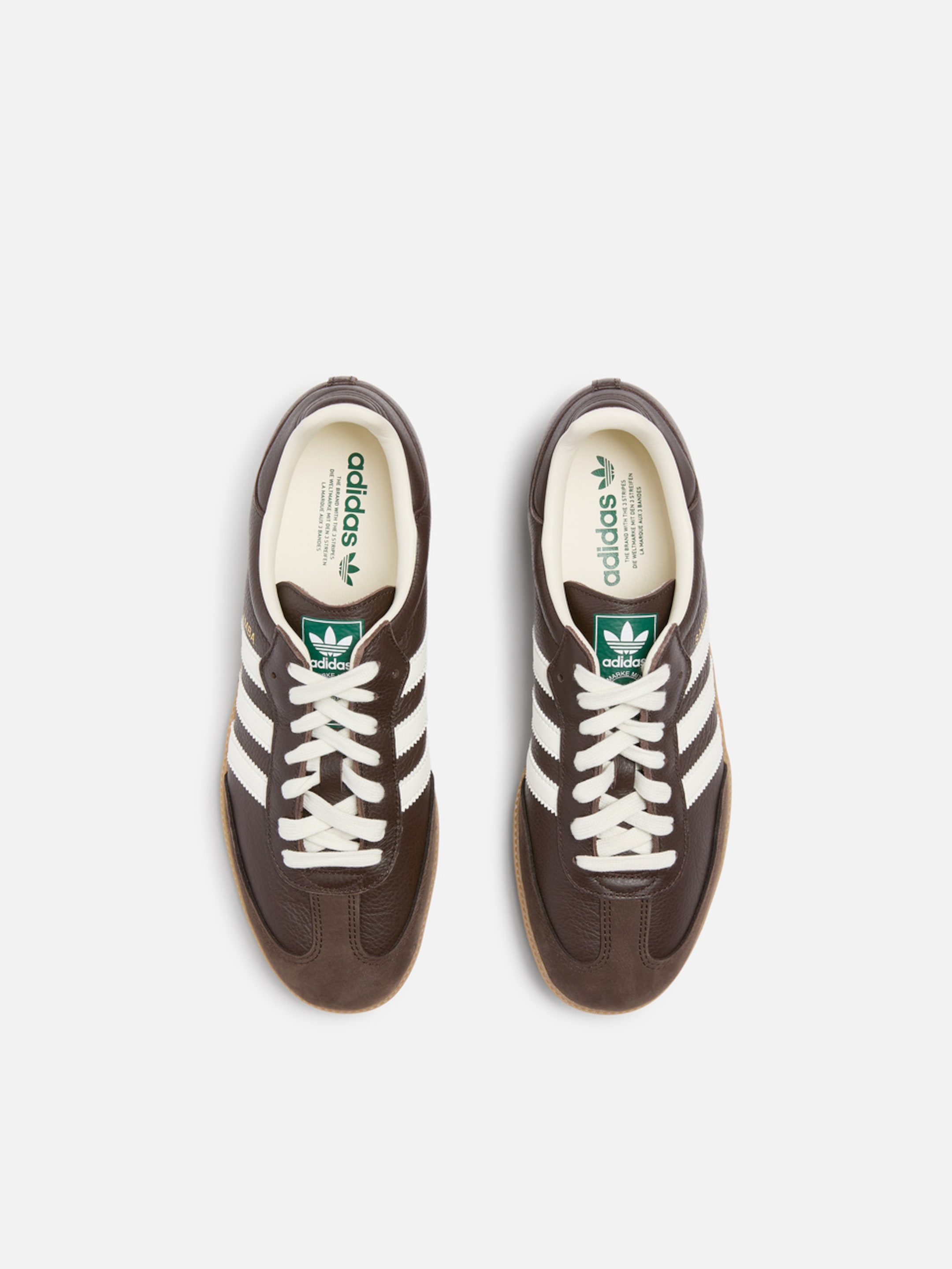 Pair of brown Adidas shoes with white stripes, white laces, and green logos, viewed from above on a white background.